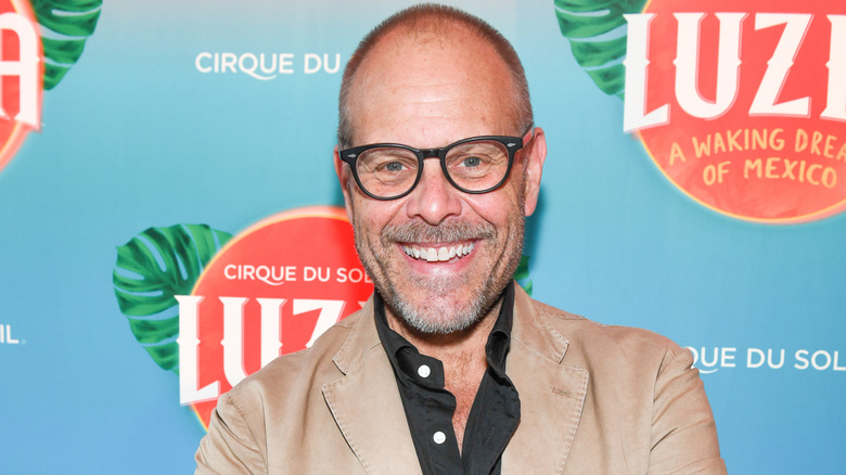 Alton Brown smiling for a picture at a red carpet event in a tan coat