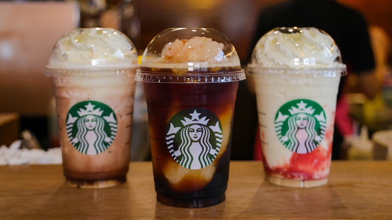 Three various Starbucks Frappuccinos on the counter in a Starbucks location with a barista and service station blurred in the background.