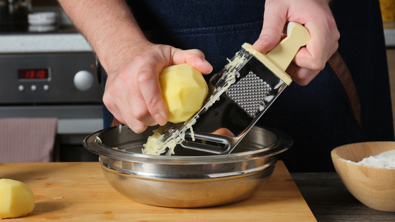 Hands grating peeled potatoes into a bowl with a box grater