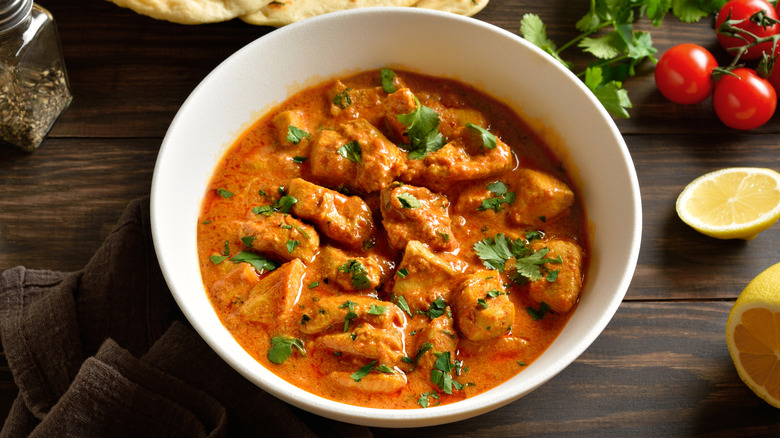 A white bowl full of butter chicken next to lemons, tomatoes, seasonings, and naan bread