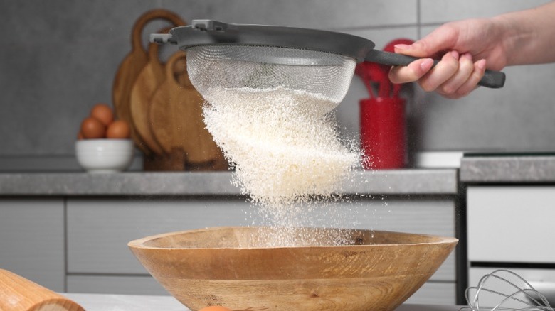 A woman sifting flour through a sieve into a wooden bowl.