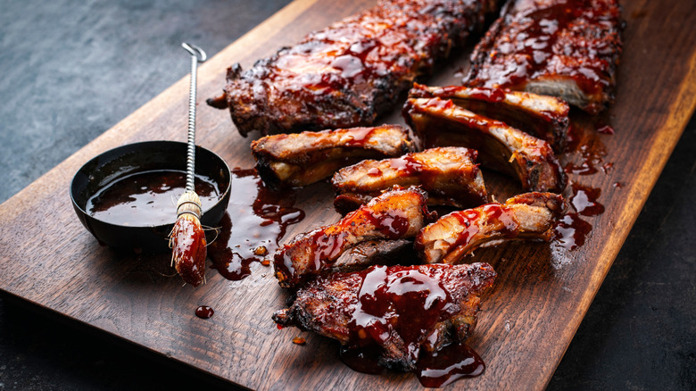 Sauce brushed pork ribs next to a rack of ribs and a bowl of sauce on a wooden plank