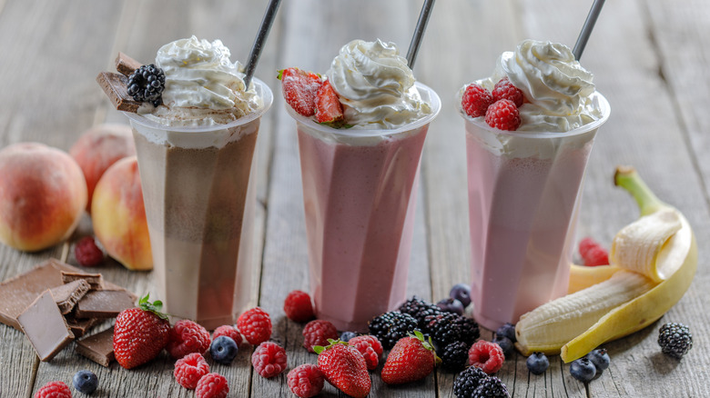 Three assorted milkshakes topped with whipped cream and fruits on a wooden surface surrounded by fruits
