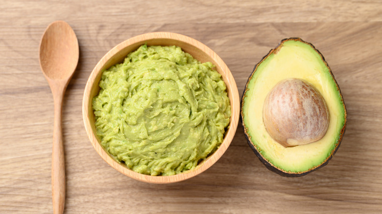 A bowl of mashed avocado next to a halved avocado and wooden spoon.