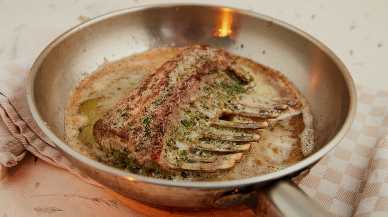 A marinated rack of lamb cooks in a stainless steel pan with a kitchen towel