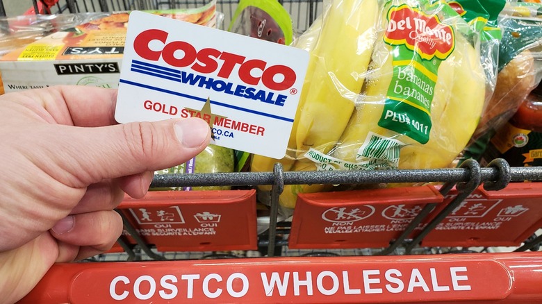 Person holding Costco membership card in front of grocery cart filled with merchandise