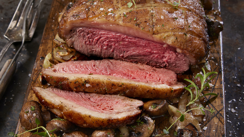 Top sirloin roast carved on a wooden board with herbs and mushrooms.