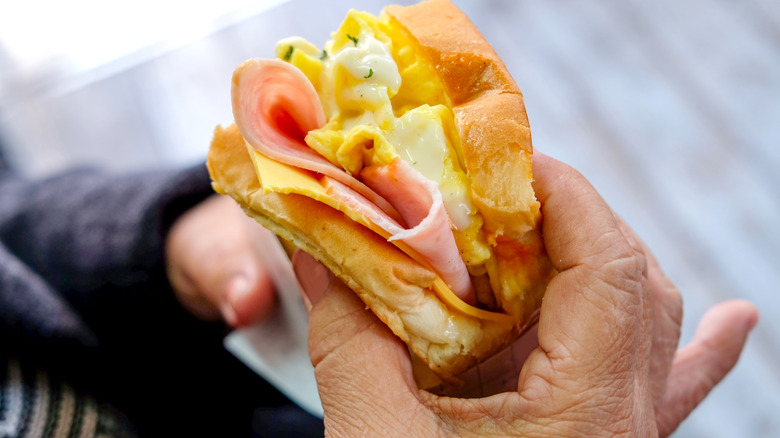 Person holding breakfast sandwich made with sliced bread, scrambled eggs, ham, and cheese