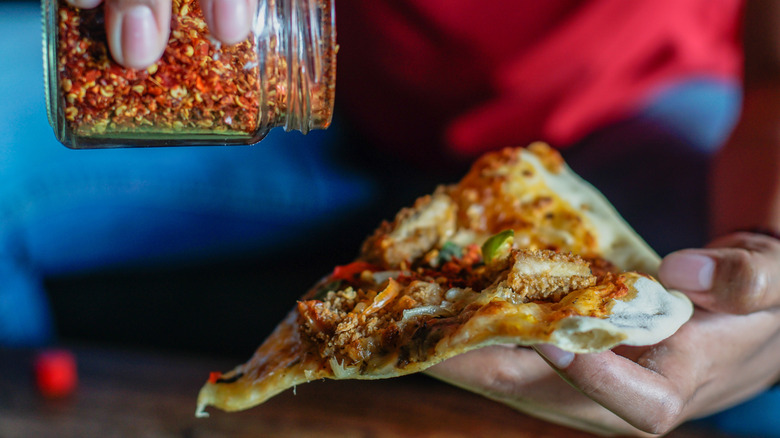 A hand adding chile flakes from a glass jar onto a slice of pizza