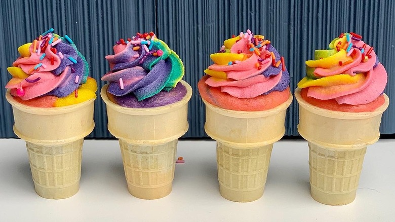 Four rainbow-colored cupcakes with frosting and sprinkles in ice cream cones