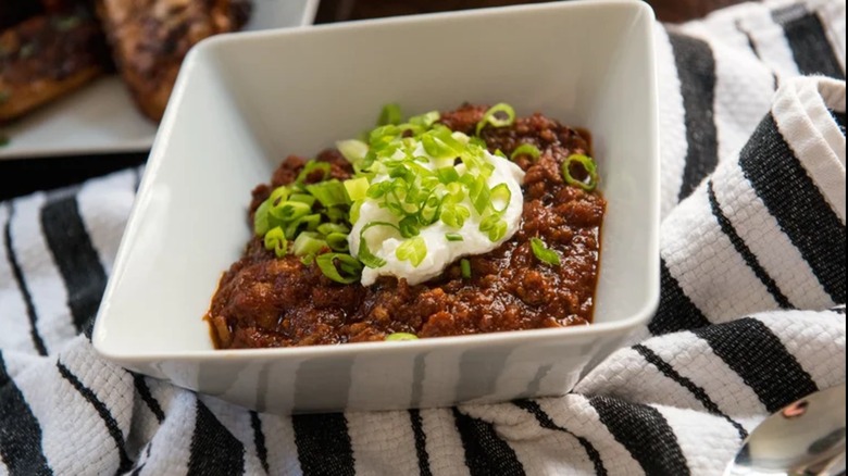 Bowl of chili on dish towel and topped with scallions and sour cream