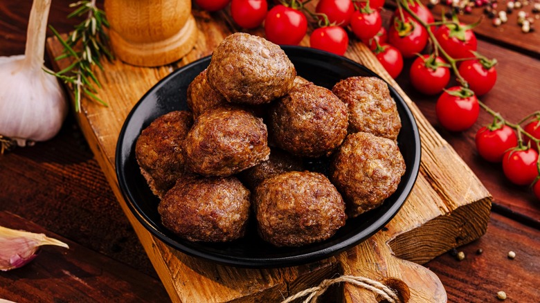 Meatballs in black bowl on wooden board beside ripe cherry tomatoes and garlic cloves
