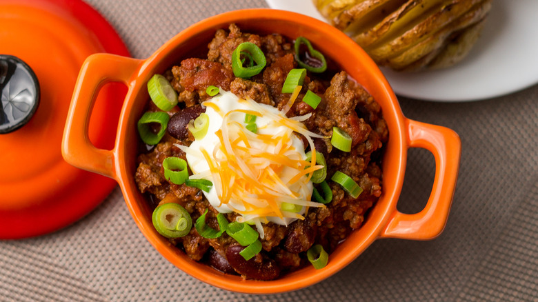 Beef and bean chili in an orange crock with sour cream, shredded cheese, and green onion