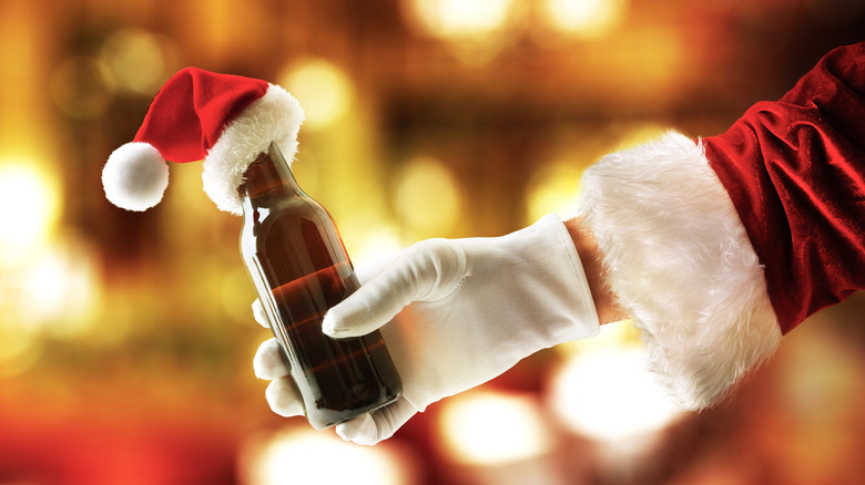 A gloved hand in a Santa coat holding a bottle of beer topped with a mini Santa hat