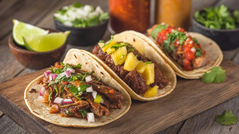 Different styles of tacos on flour tortillas with taco garnishes in the background