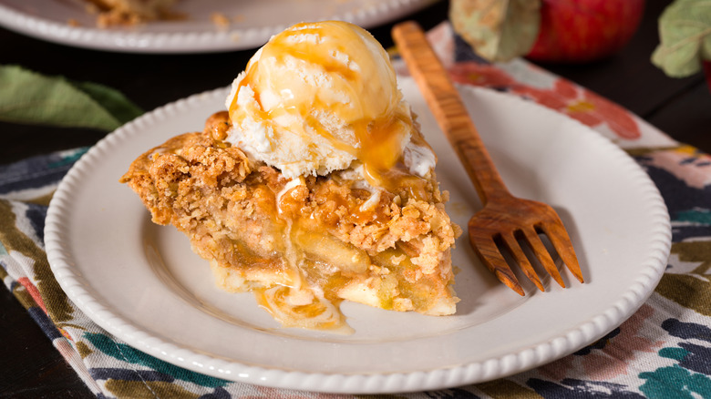 A slice of apple pie a la mode on a plate in front of apples and a pie