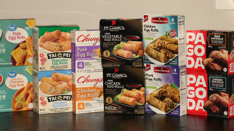 An array of six brands of frozen egg rolls in their packaging and side by side on a table