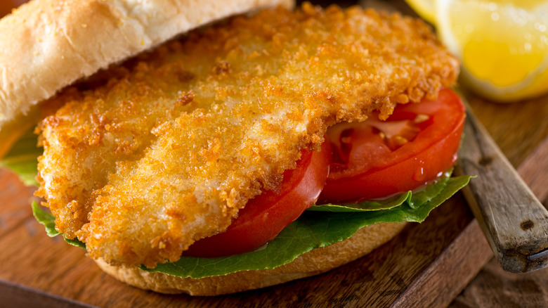 A fried fish sandwich with lettuce and tomatoes with lemons in the background.