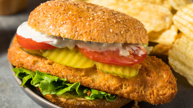 A fried fish sandwich with tomatoes and pickles next to a plate of chips.