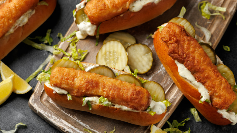 Fish sticks in hot dog buns with pickles, sauce, and lettuce on a cutting board and dark surface