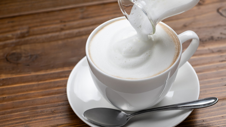 white coffee mug sitting on top of saucer with metal spoon and froth being poured on top