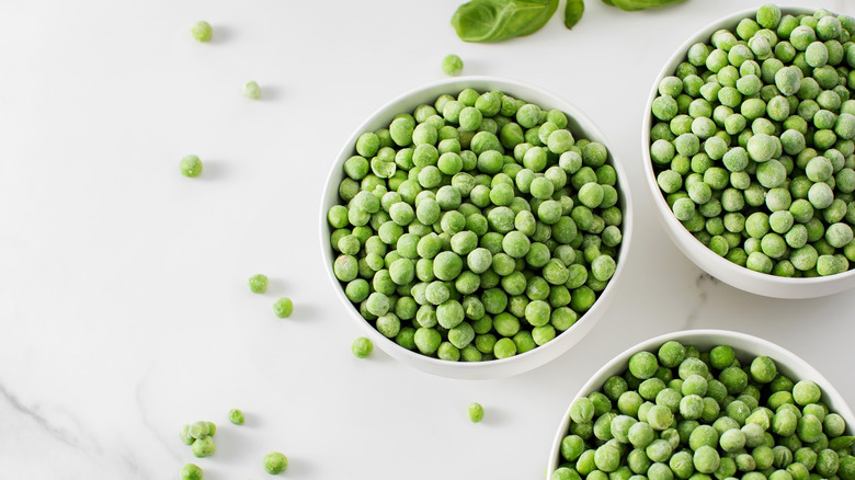 Three bowls of frozen peas on a white surface littered with more peas and some basil leaves