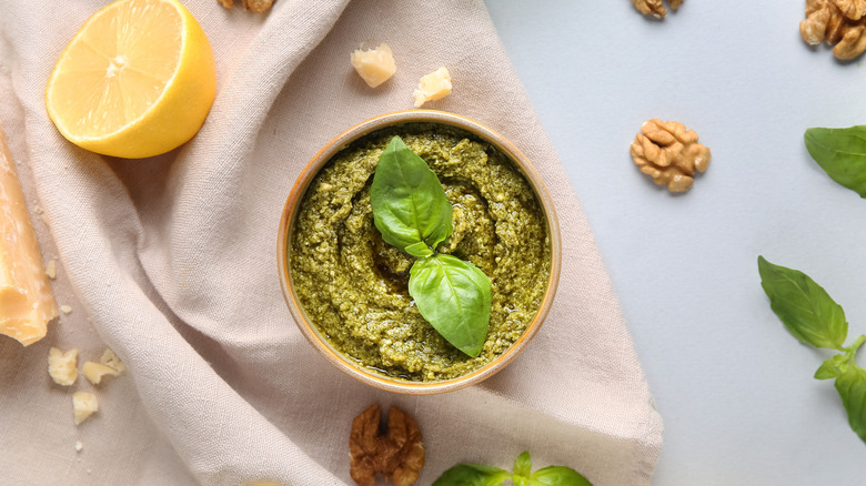 A bowl of pesto topped with basil on a white cloth, surrounded by parmesan cheese, walnuts, a halved lemon, and more basil leaves