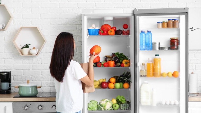 Young woman putting groceries into open fridge in kitchen, back view