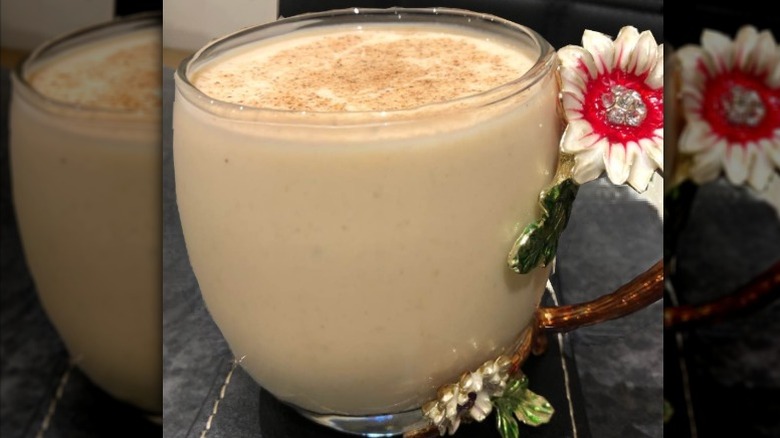 a glass cup with a flower handle filled with creamy banana punch with cinnamon and nutmeg on top