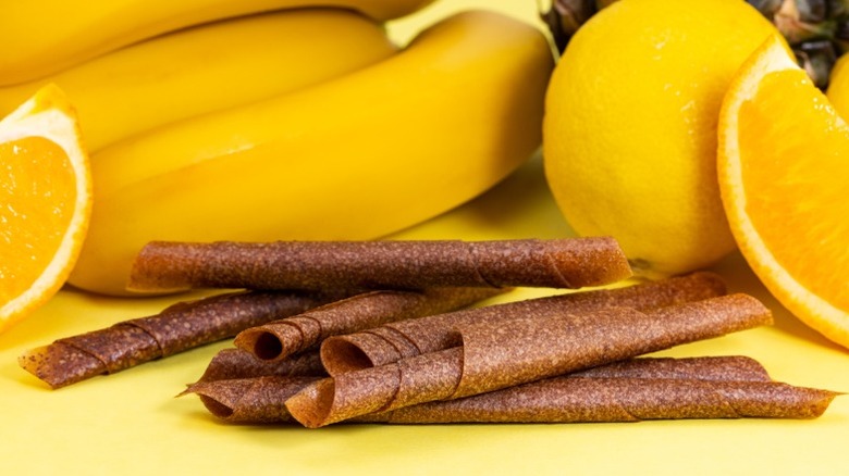 rolls of fruit leather on a yellow background surrounded by bananas, lemons, and oranges