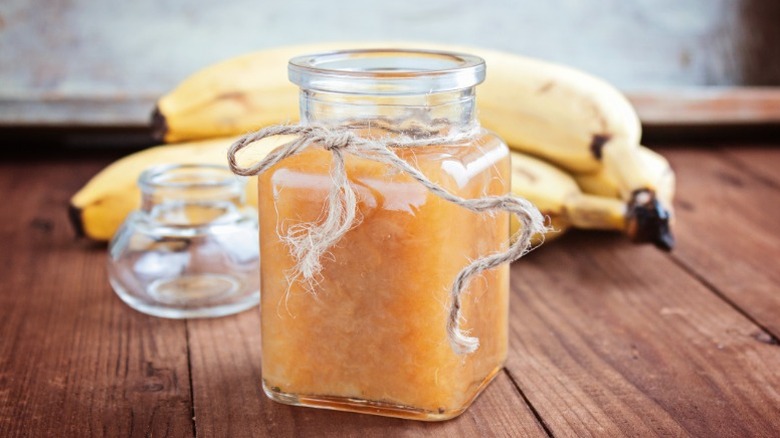 a glass jar filled with banana jam and tied with twine on a wooden counter with bananas behind it