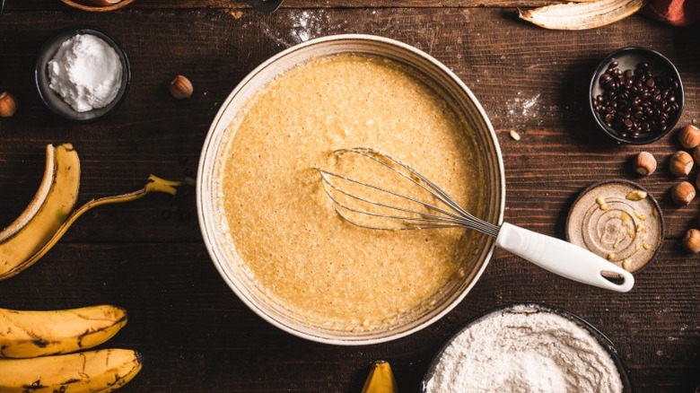 A bowl of batter with a whisk on a counter surrounded by ingredients like bananas, flour, nuts, and coffee beans