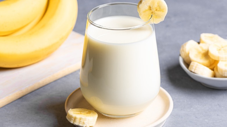 a glass of banana milk next to a plate with sliced bananas and a bunch of whole bananas