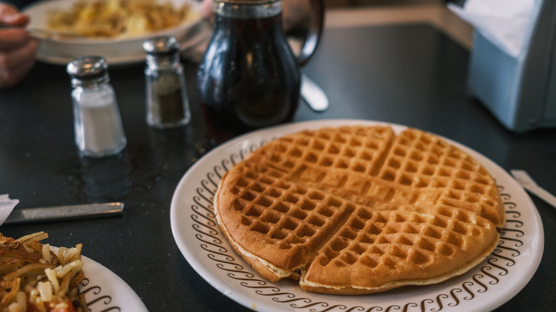 A large Waffle House waffle on a white plate beside syrup, salt, pepper, and other plates of food.