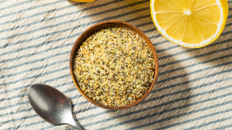 A small wooden bowl containing lemon pepper seasoning on a striped cloth with a spoon and a halved lemon beside it