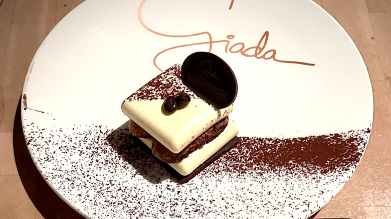 White plate reading "Giada" at the top in cursive with a gourmet piece of tiramisu in the center and cocoa powder dusted across the plate