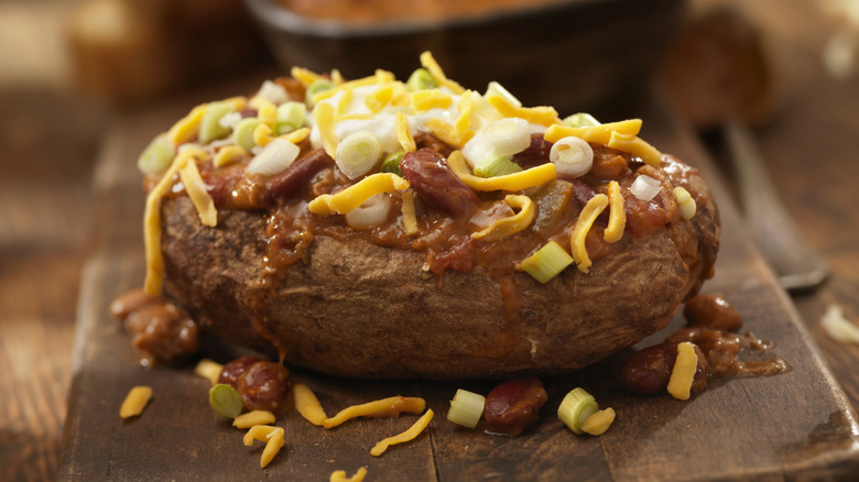A baked potato topped with chili, cheese, green onion, and sour cream, sitting on a cutting board