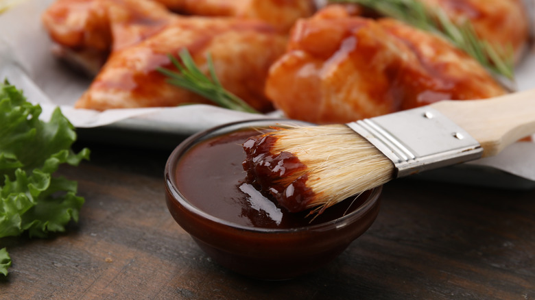 A closeup of a bowl of BBQ sauce with a brush with chicken behind