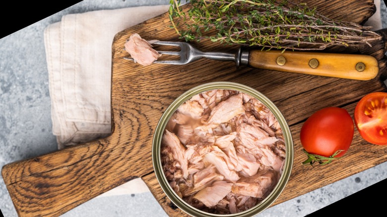 Tinned tuna on a wooden serving board with herbs, tomatoes, and a fork