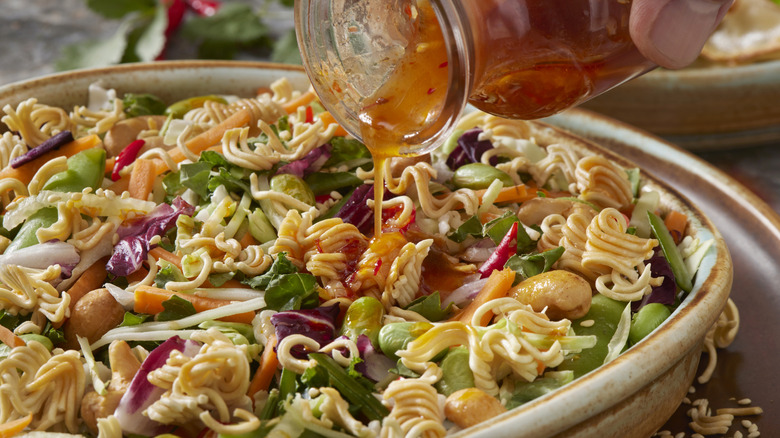 Bottle of sweet and sour sauce dressing being drizzled over ramen noodle slaw with cabbage, carrots, edamame, and cashews