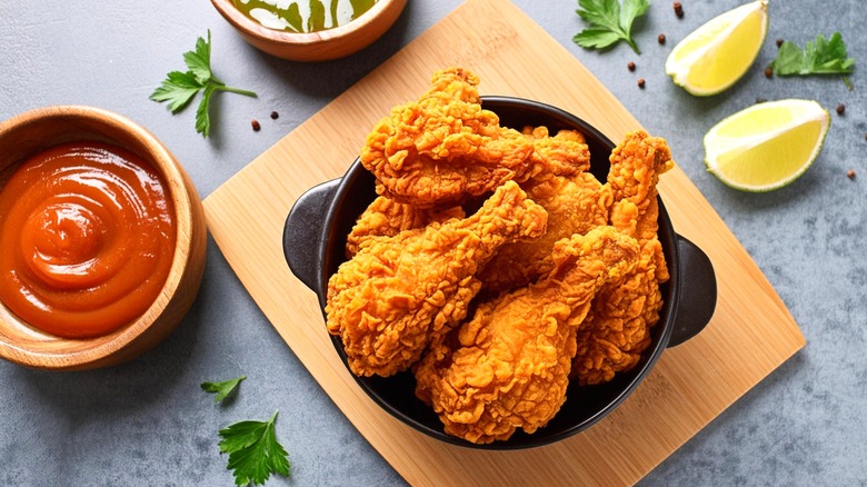 Pieces of golden fried chicken in a black bowl on a wooden board with cut lime quarters and a small bowl or red sauce next to it