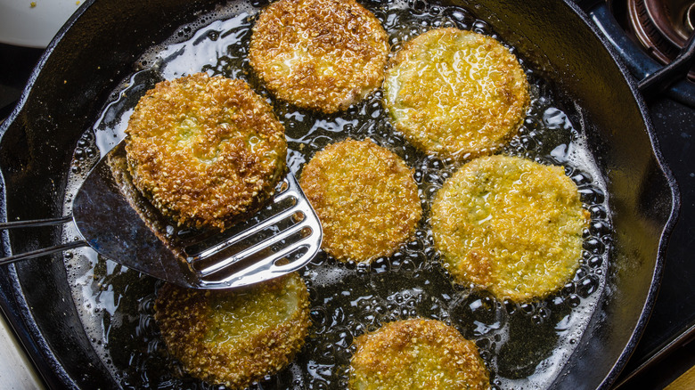 Spatula scooping one of several fried green tomatoes bubbling in a cast iron pan.