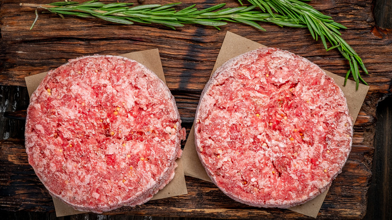 Frozen raw beef burger patties on wooden cutting board with sprigs of rosemary