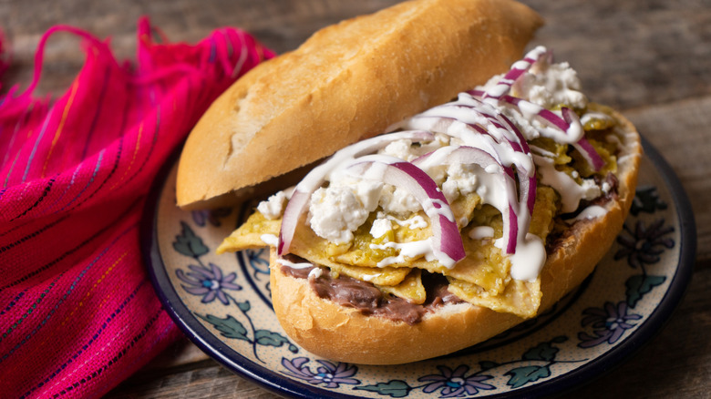 A refried bean and chilaquiles verdes torta on a bolillo roll with queso fresco, red onion, and crema
