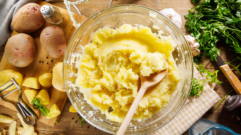 Preparing mashed potatoes at home in a glass bowl with a peeler, cutting board with boiled potatoes, pepper, parsley, garlic, mixing wooden spoon