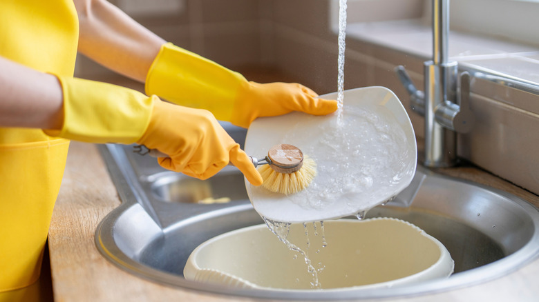 A woman washes the dishes in kitchen sink while wearing yellow gloves and apron, and using a brush to clean one plate.