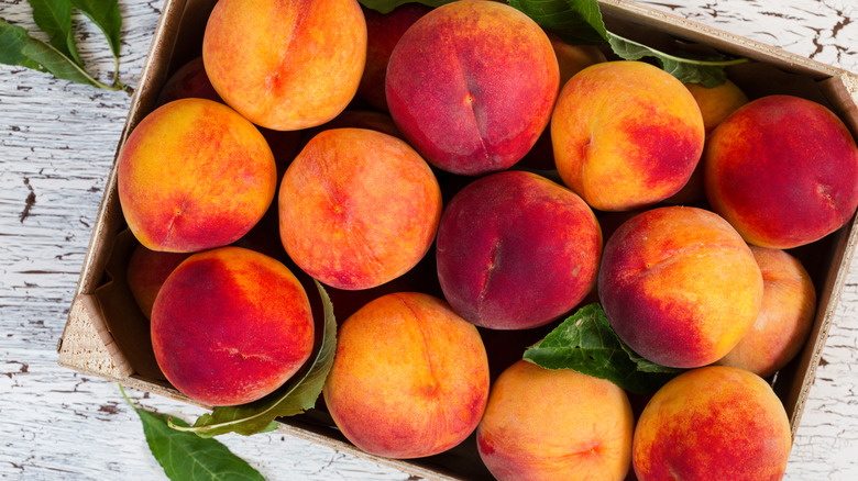 Box of fresh ripe peaches surrounded by green leaves in a wooden crate.