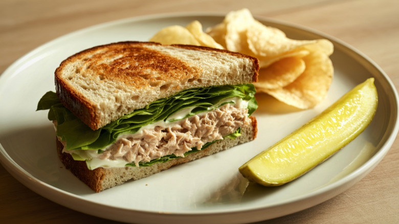 A tuna salad sandwich on toasted bread rests on a plate next to a pickle spear and potato chips