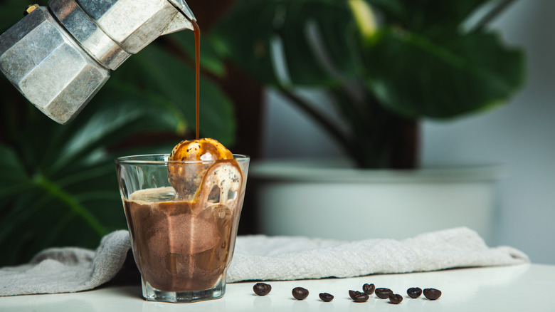 hand pouring coffee over cup of ice cream next to coffee beans