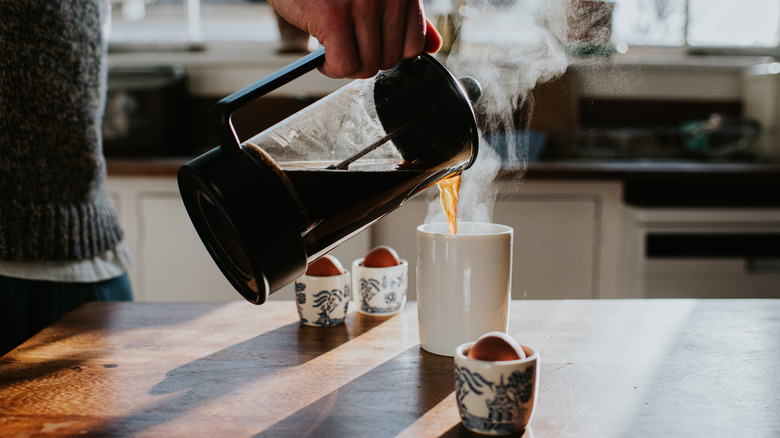 A person pouring hot coffee into a white mug on their counter